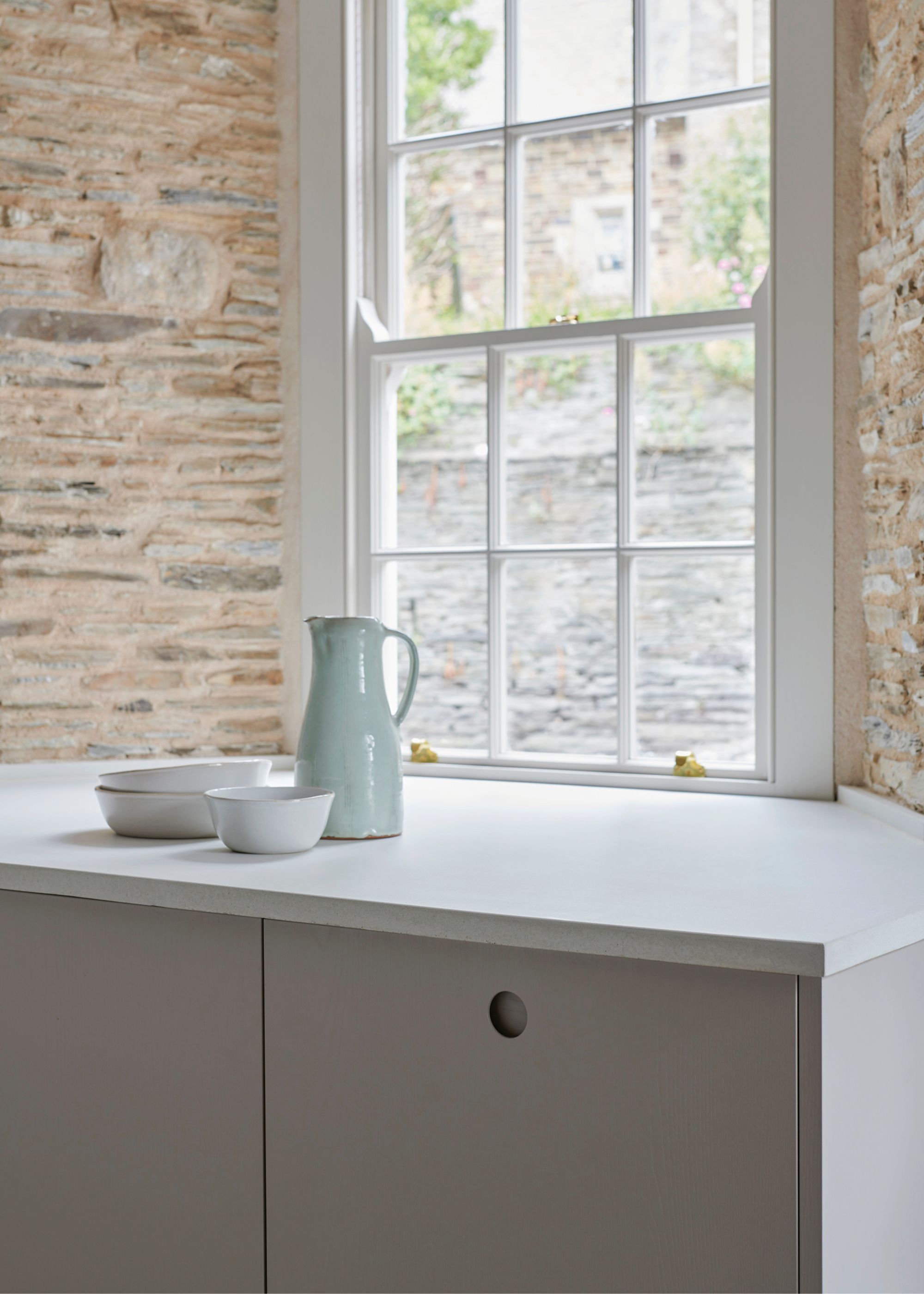 A bright kitchen with white concrete worktops and a bowl as well as water dispenser placed on top of it beside a large kitchen window