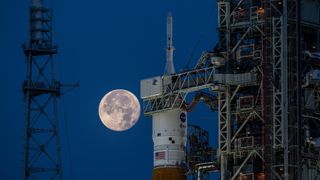 the full moon hangs in frame with the orion, inside its payload shell atop the SLS rocket.