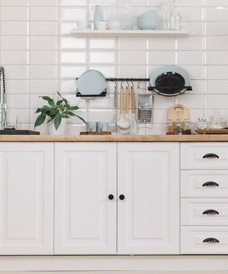 An image of white kitchen cabinets and a wooden countertop with plates and utensils on a metal rack in the background