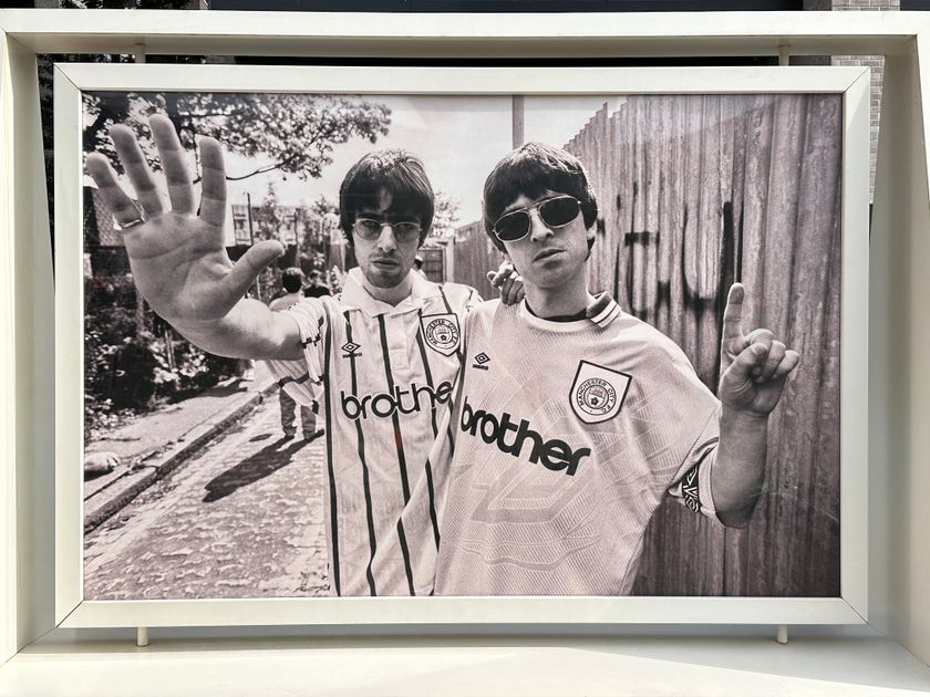 A black-and-white photo of Oasis band members Liam and Noel Gallagher wearing Manchester City football jerseys. Liam holds his hand up, palm facing out, while Noel holds his hand up with his index finger pointing to the sky.