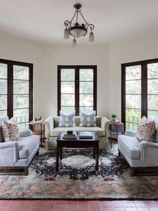 sunroom with angled white wall and windows all round with white sofas and blue cushions and dark wood tables on oriental rug with terracotta floor tiles
