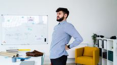 A man in a shirt stands in an office, leaning forward slightly, with his left hand pressed against his lower back. In front of him is a messy desk, behind him we see a whiteboard, shelving and an arm chair.
