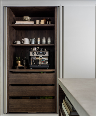 A hidden coffee bar with a silver coffee maker and wooden shelves.