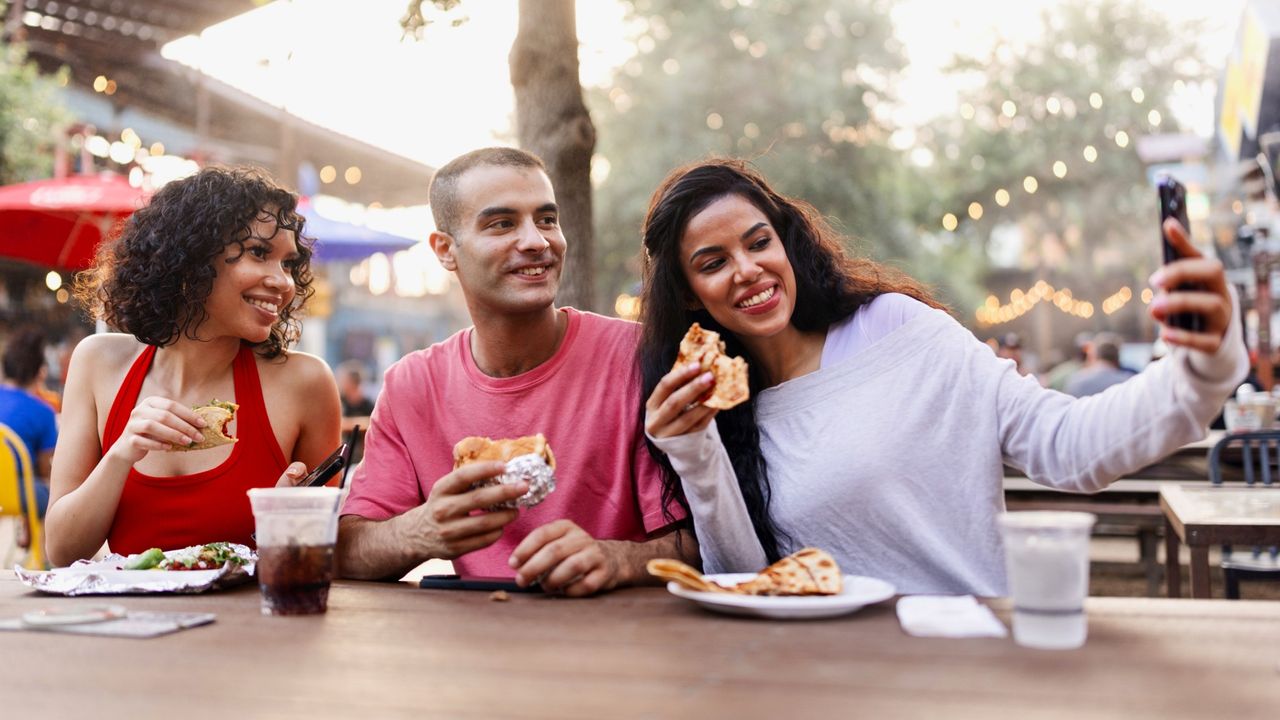 A stock photo of friends sitting around a table at a food festival, taking a selfie.