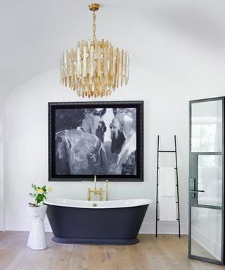 bathroom with white walls, wooden floor, black rolltop tub, statement monchrome artwork, dramatic gold chandelier