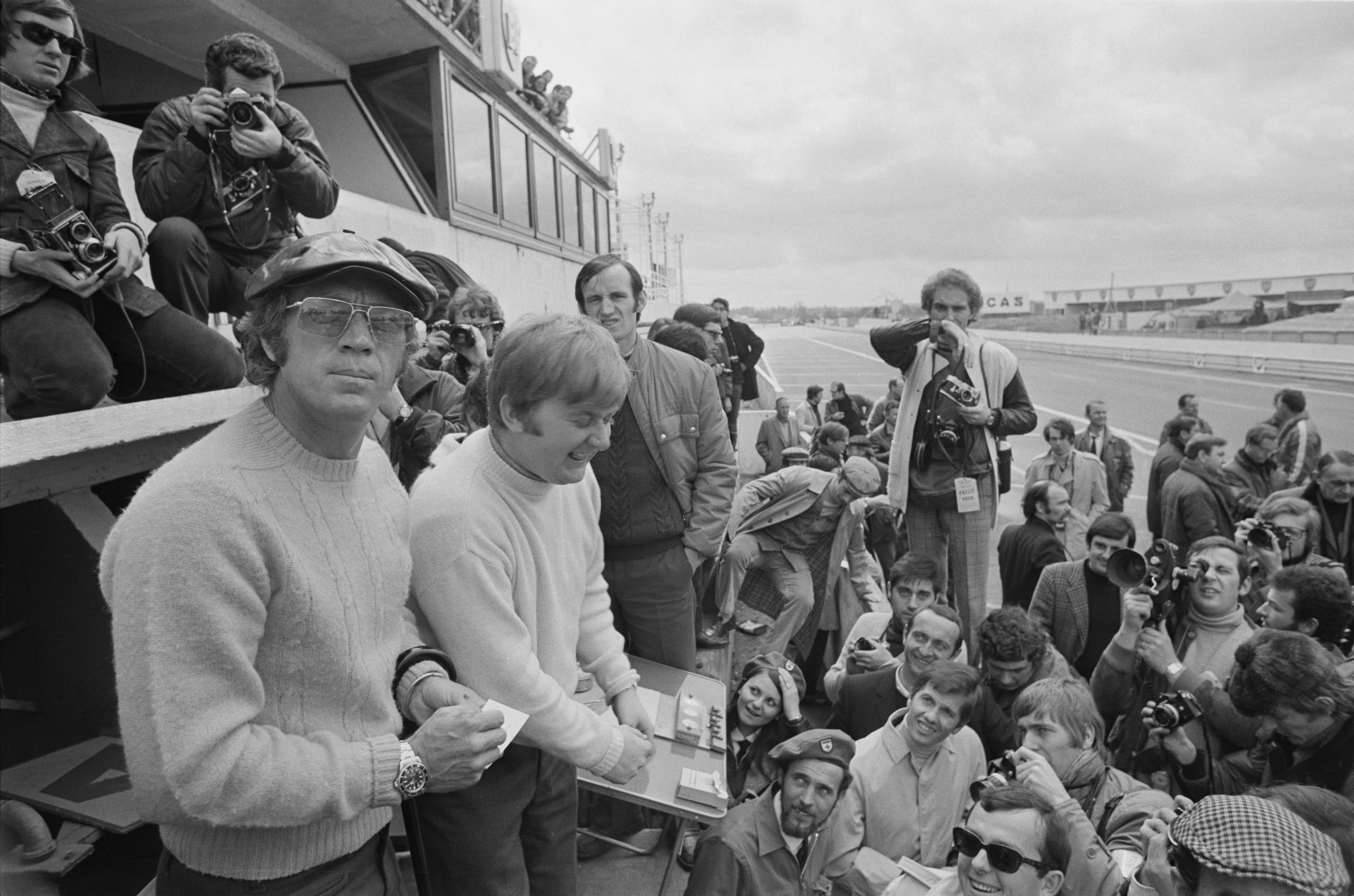 Steve McQueen signing autographs at Le Mans. He is wearing 1970s style sunglasses and a cream cable knit jumper