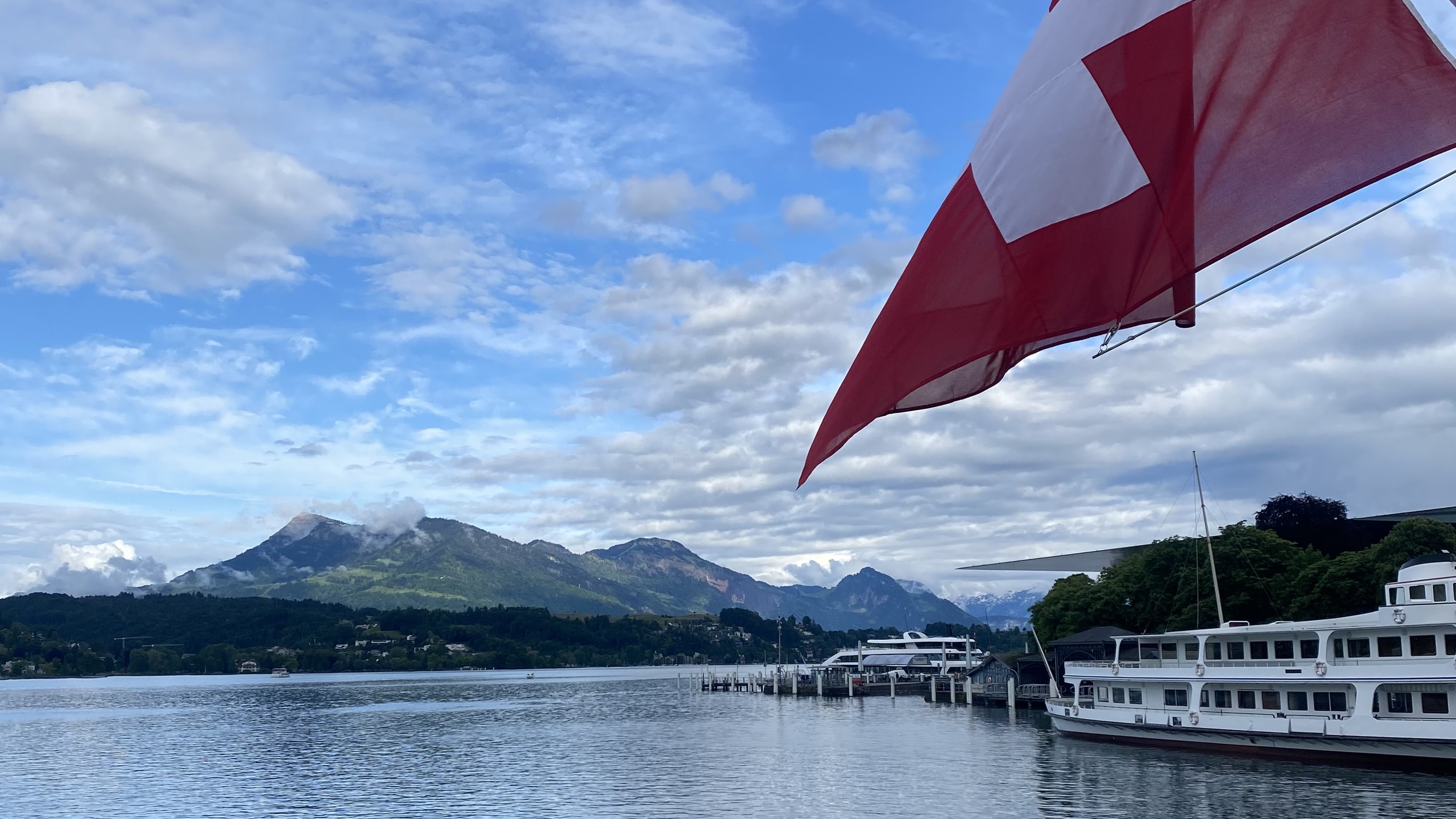 The Swiss flag flies over Lake Lucerne, with mountains in the distance, on a clear day.