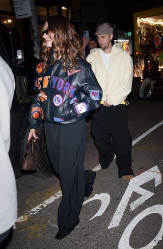 A photo of Hailey Bieber wearing a black leather bomber jacket covered in New York Knicks patches, loose black trousers, and black Ferragamo loafers.