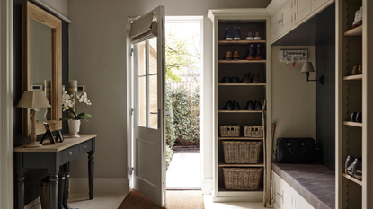a back entryway mudroom with built-in shelving, several baskets for storage, a built-in bench, and a small table holding a lamp, flowers and picture frames to the left