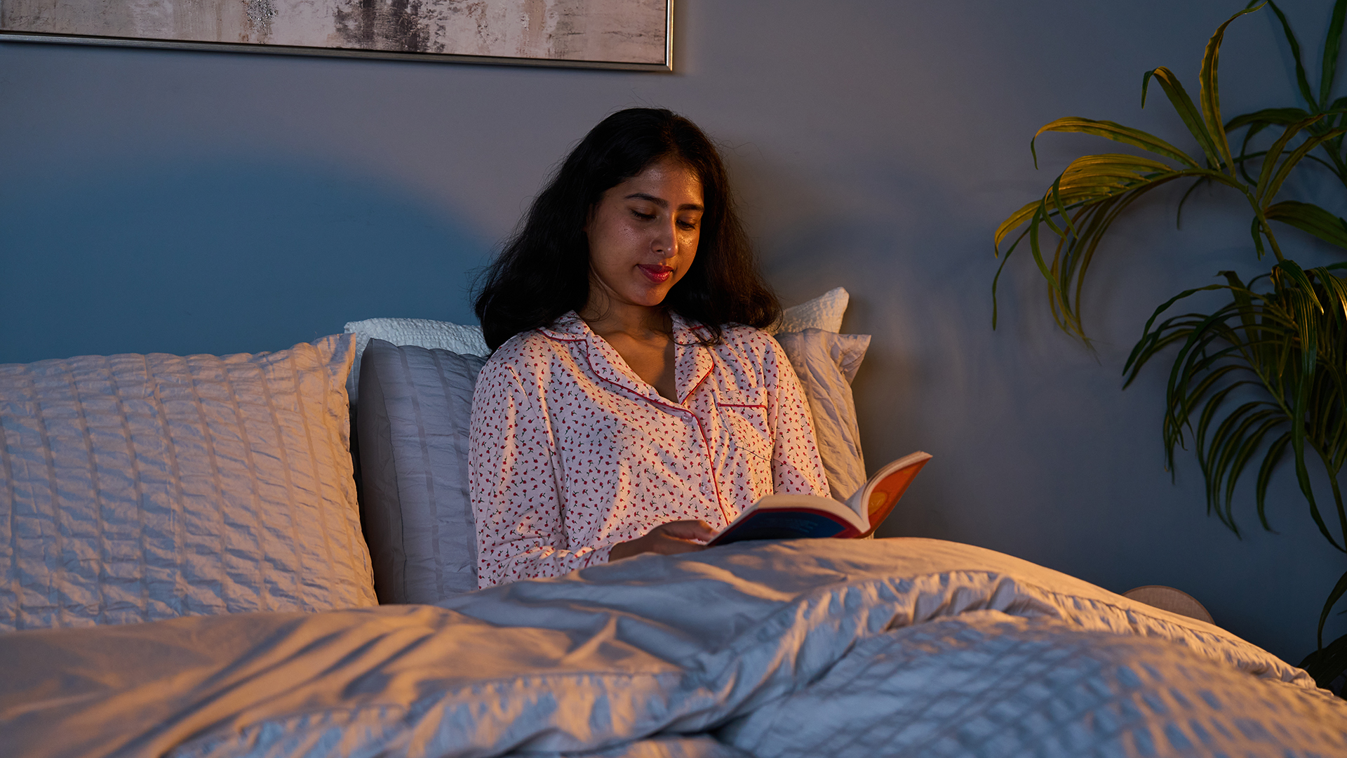 A woman sits up in bed reading a book in dim lighting