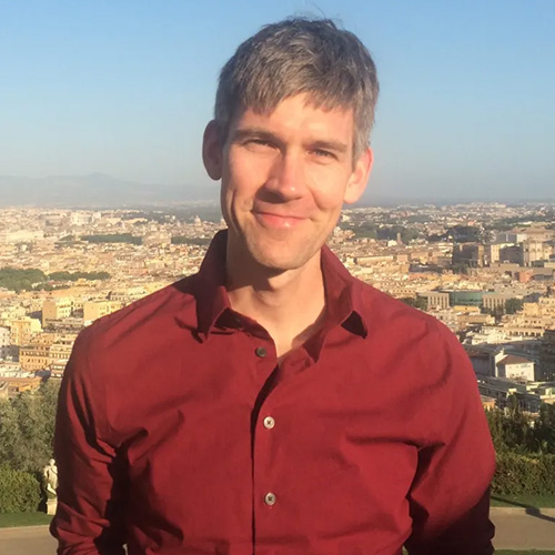 A gray-haired man wearing a red button-up shirt smiles at the camera