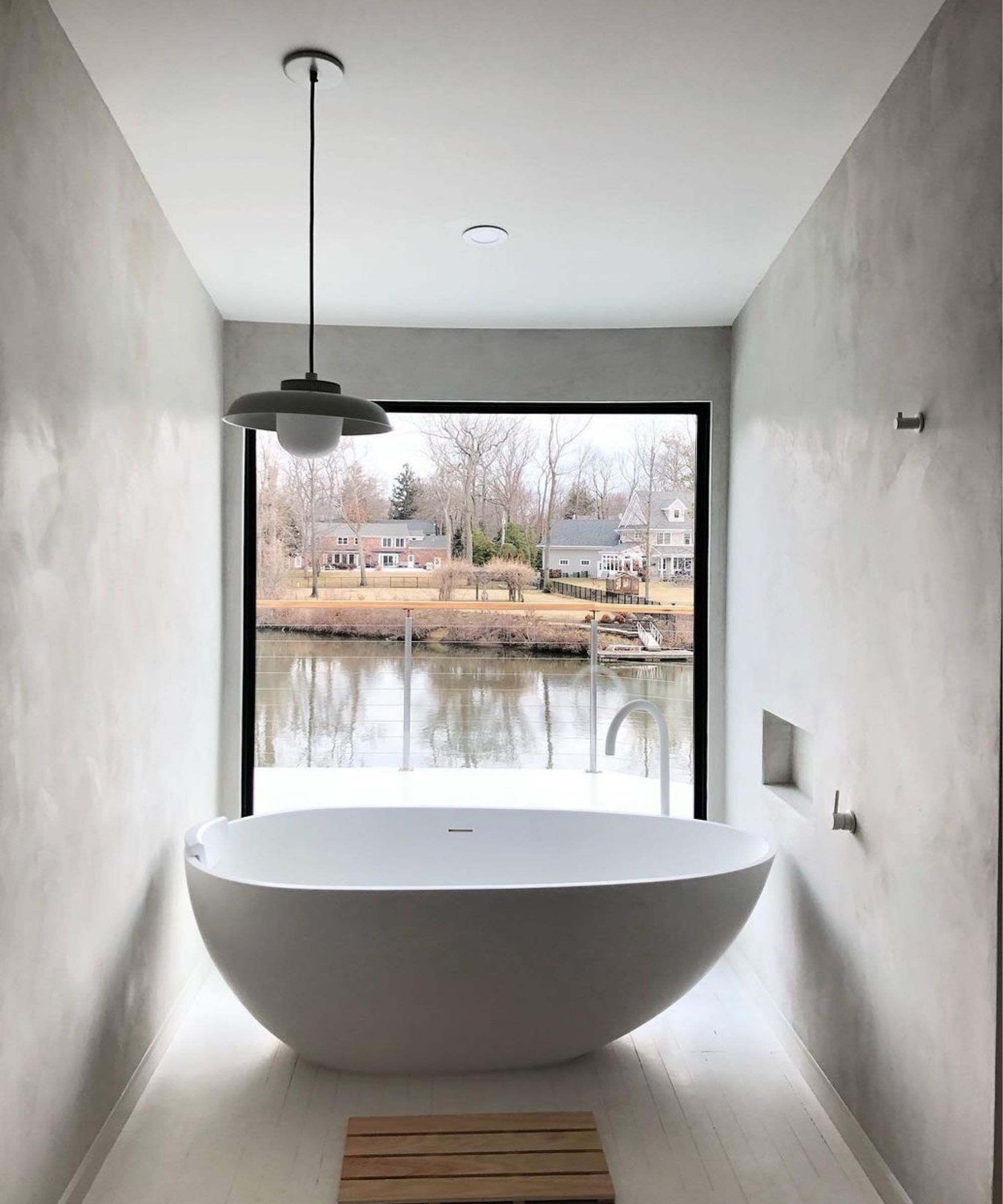 A minimalist bathroom with smooth grey plaster walls and white painted floorboards, centred on a large white oval freestanding bath positioned directly in front of a full-width black-framed picture window overlooking a calm river and houses beyond. A single grey pendant light hangs above and a slatted wood bath mat sits below.
