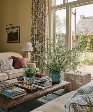 english country decorated living room with soft yellow walls and a leather ottoman decorated with a large foliage display by french doors with floral curtains