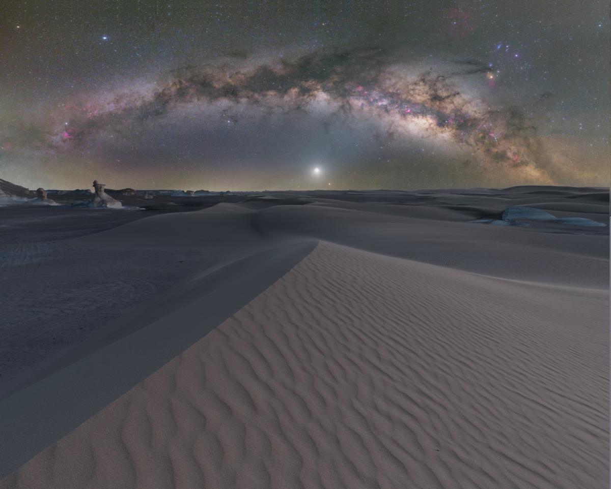 A wavy desert sand dune sits beneath a bright Venus, hung low in the sky with the arch of the Milky Way curved overhead.