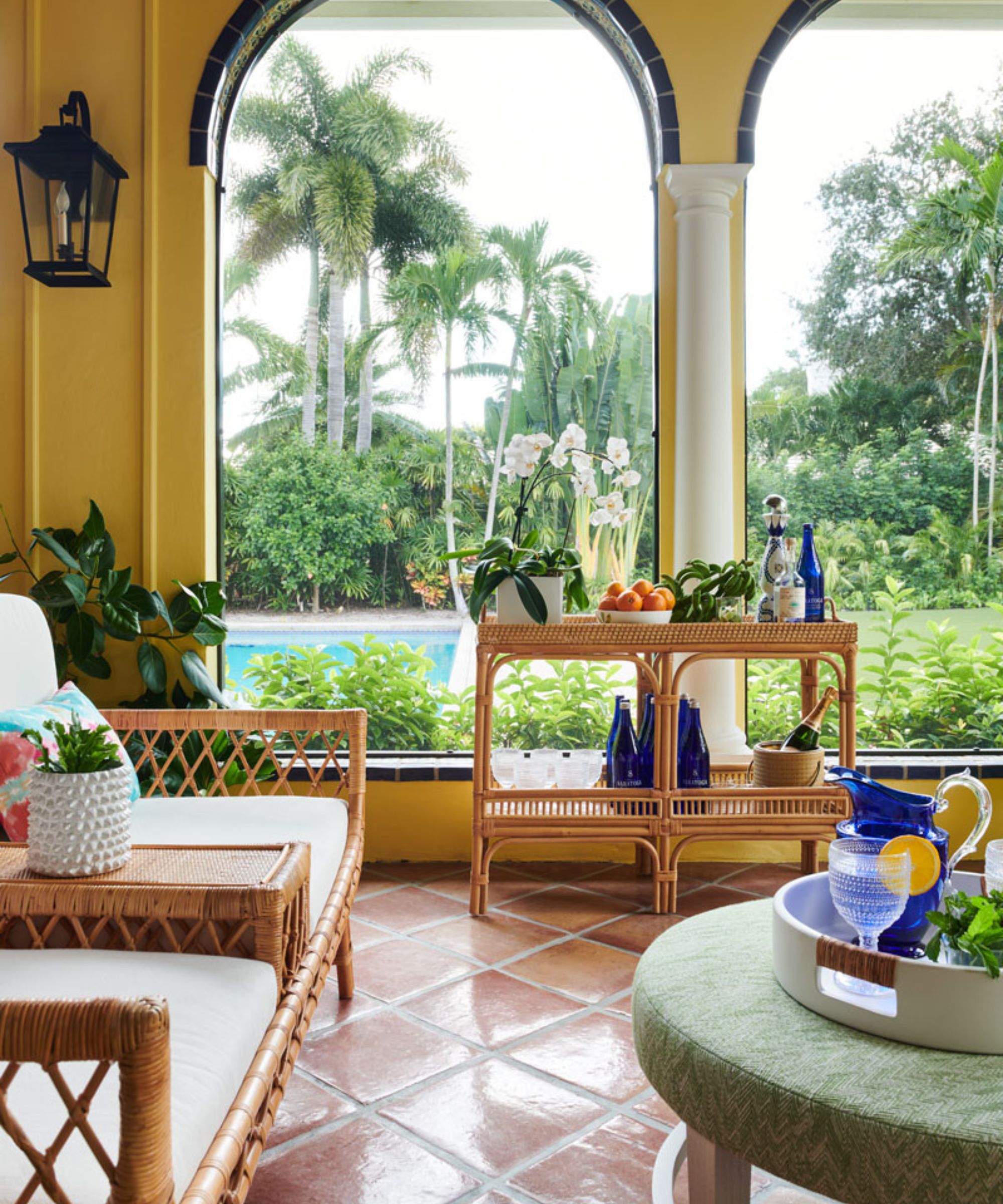 Porch with terracotta tiled floor, yellow painted arched walls, rattan bar cart with drinks and plants, rattan lounge chairs, and a green ottoman with a tray holding glasses and a blue pitcher