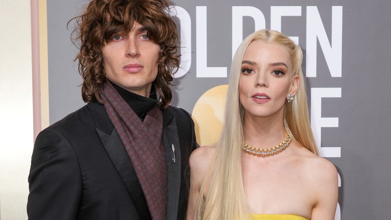 Anya Taylor-Joy wears a strapless yellow top and matching skirt and has long blonde hair while her husband Malcolm McRae wears a dark suit jacket with a brown scarf