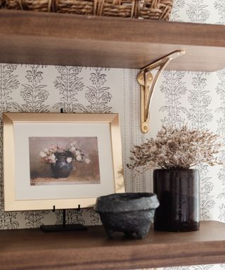 Close up of a wooden wall shelf with a gold framed picture and a black vase of dried small flowers. Another shelf above with the bottom of a woven basket visible