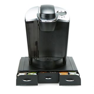 A black coffee pod machines sits on top of a black storage box with three dividing drawers