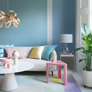 living room with blue walls, white couch with cushions, a run with blue, yellow, purple and brown patter, a pink side table, a white coffee table and a fancy brown ceiling pendant light