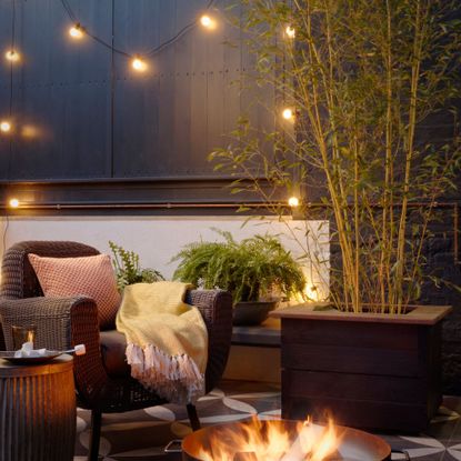 outdoor seating area with rattan chair in front of a firepit, with festoon lights on the fence behind