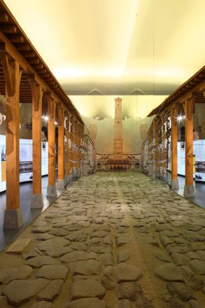 View of the Sala Forma Urbis, ancient stone floor, wooden framed pillars, exhibition pieces on either side, monument and wall lighting at the end of the pathway