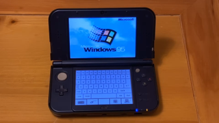 A black, New-style Nintendo 3DS sits on a wooden table. The lower screen shows the handheld console's touchscreen keyboard. The upper clamshell screen show the Windows 95 startup logo.