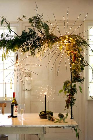 White rustic dining room with a wooden white table. The table has a table rod on it decorated with garland lights and foliage.