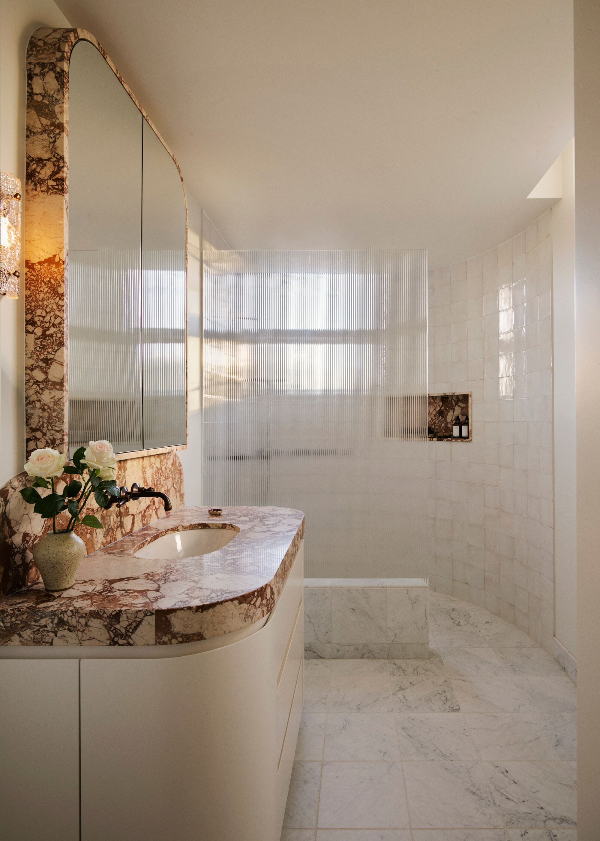 A white bathroom with a walk in shower, a marble counter, a vase of roses, and ribbed glass