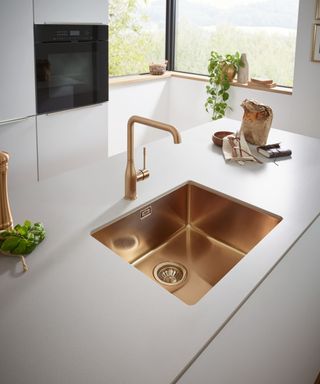 A contemporary kitchen with a gold undermount sink and matching tap. A light grey stone worktop, sleek cabinetry, and a large window create a bright space.