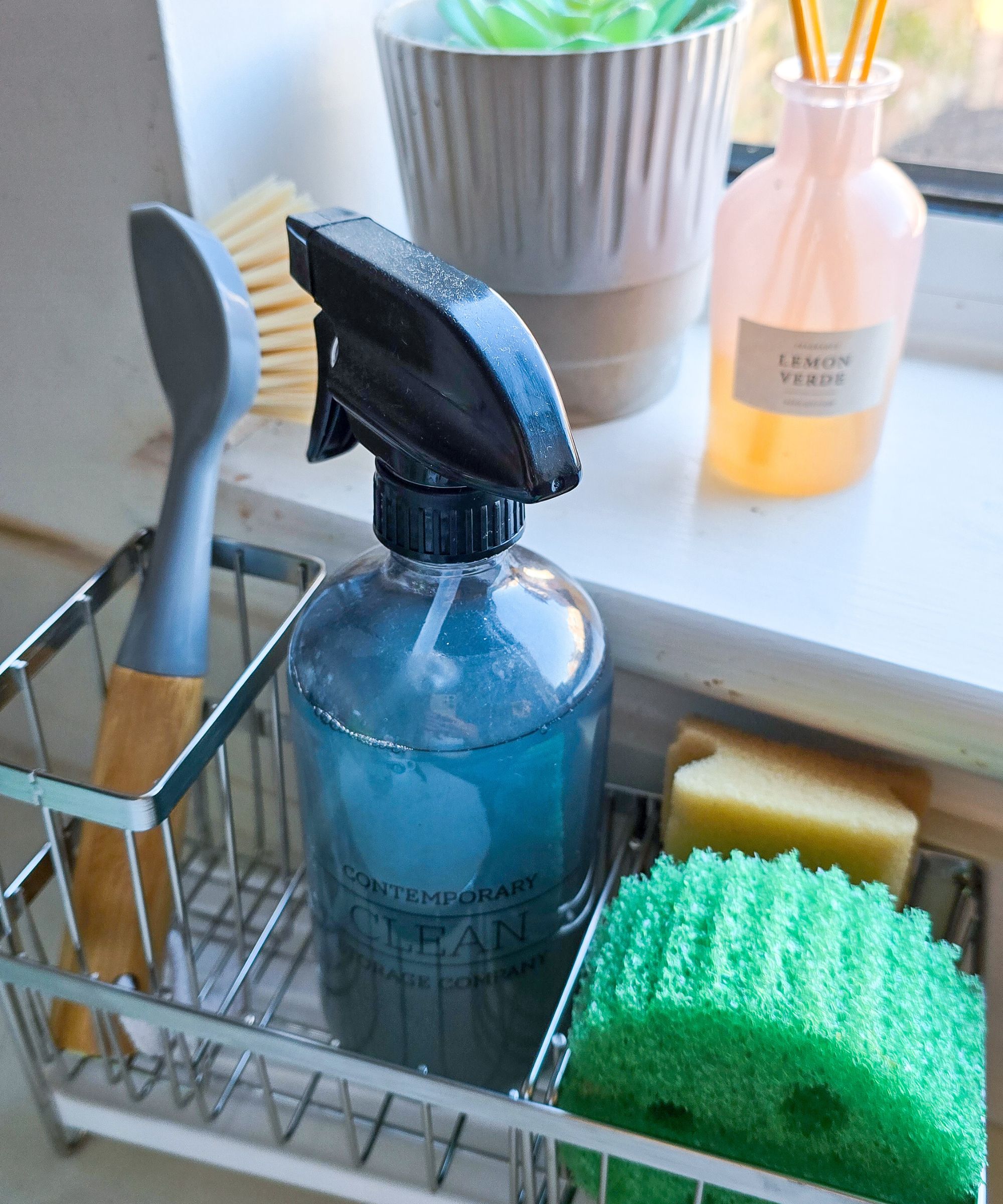 A kitchen sink caddy with a brush, a glass bottle, and two sponges in it, with a windowsill with a plant and diffuser behind it