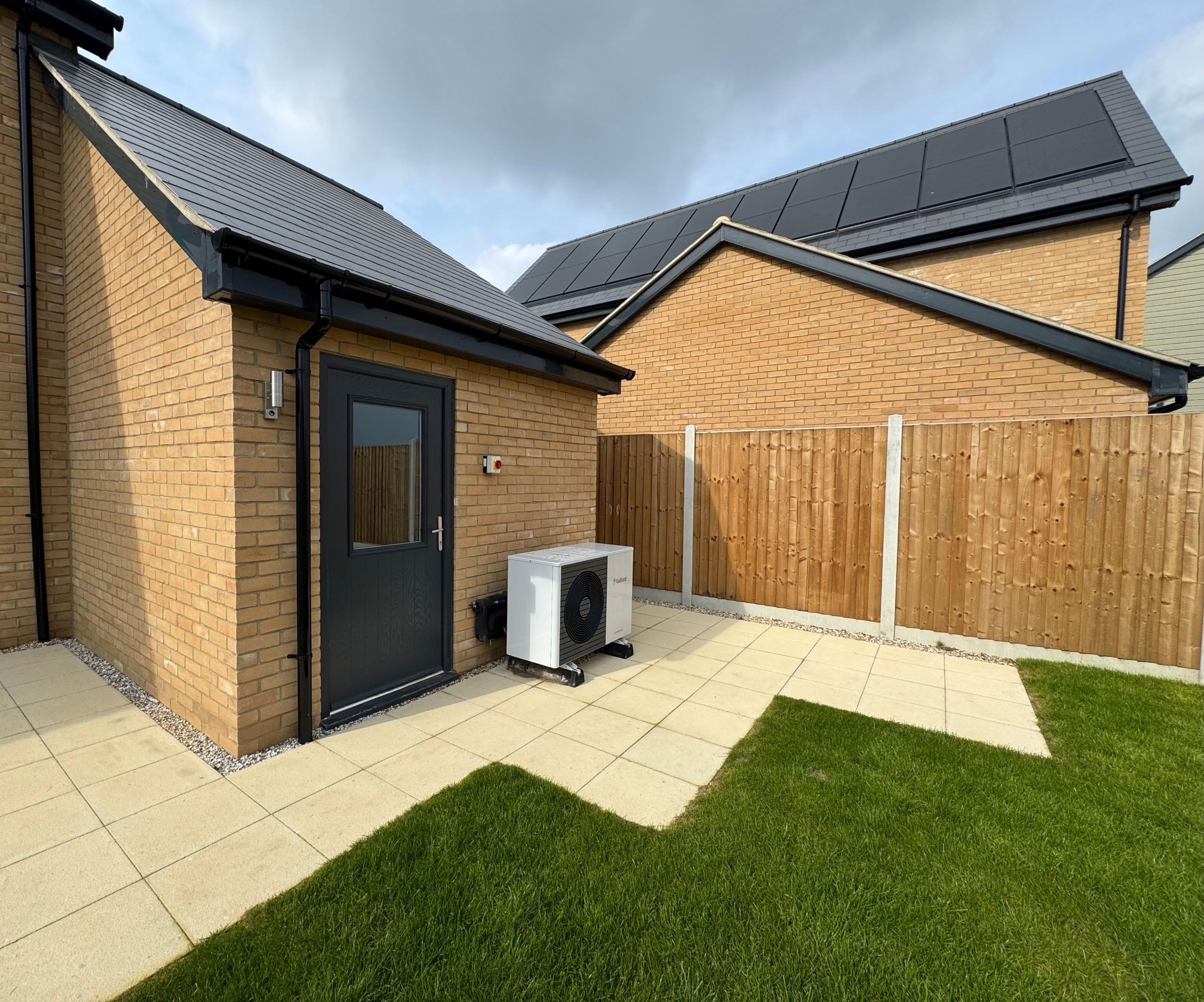 A buff brick house with a lush green lawn and a buff coloured patio and a heat pump beside a grey door
