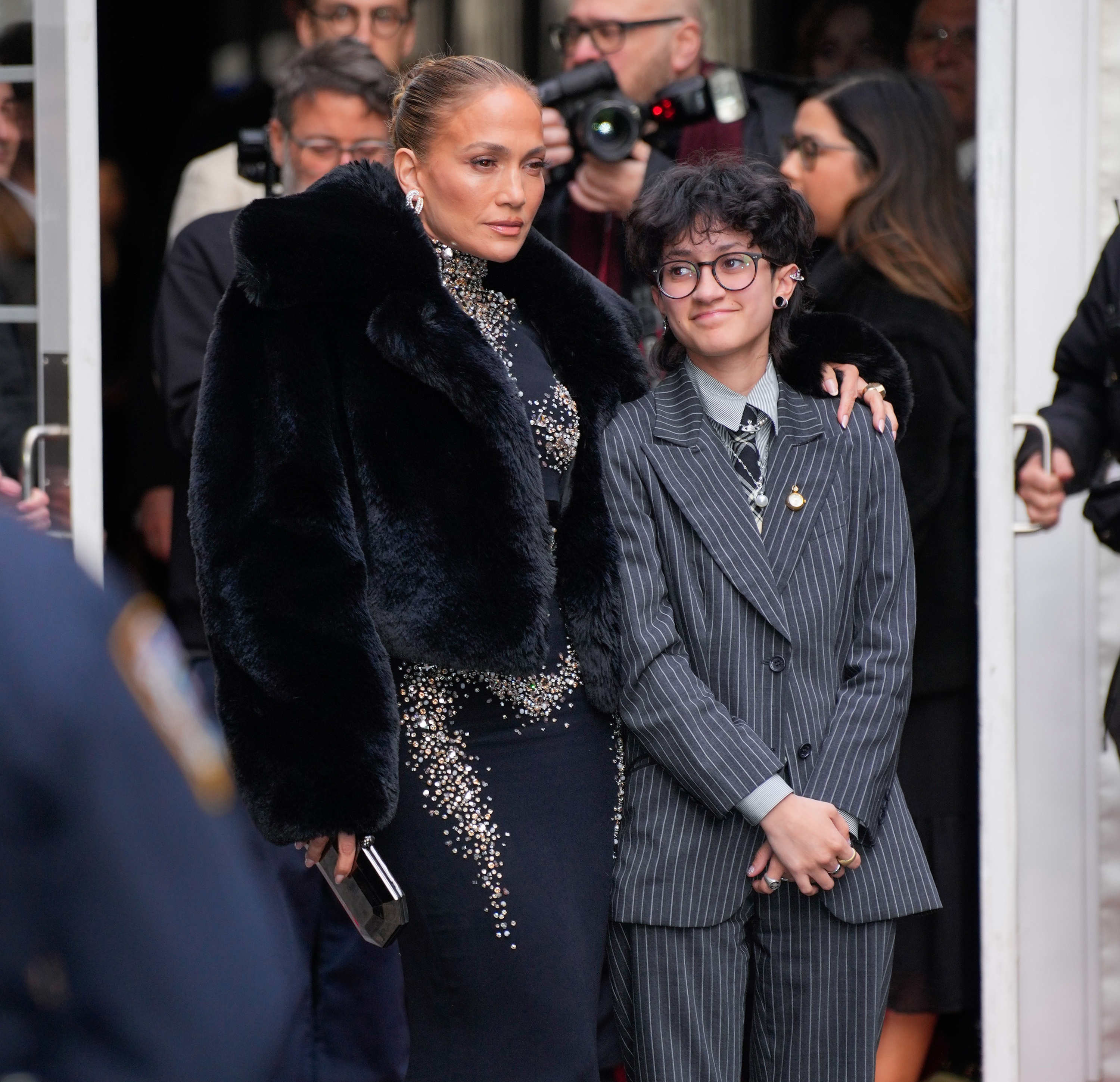 Jennifer Lopez in a black embellished gown with a black fury coat and Emme Muñiz in a gray pinstriped suit at "Othello" opening night at the Barrymore Theater in Midtown on March 23, 2025 in New York City