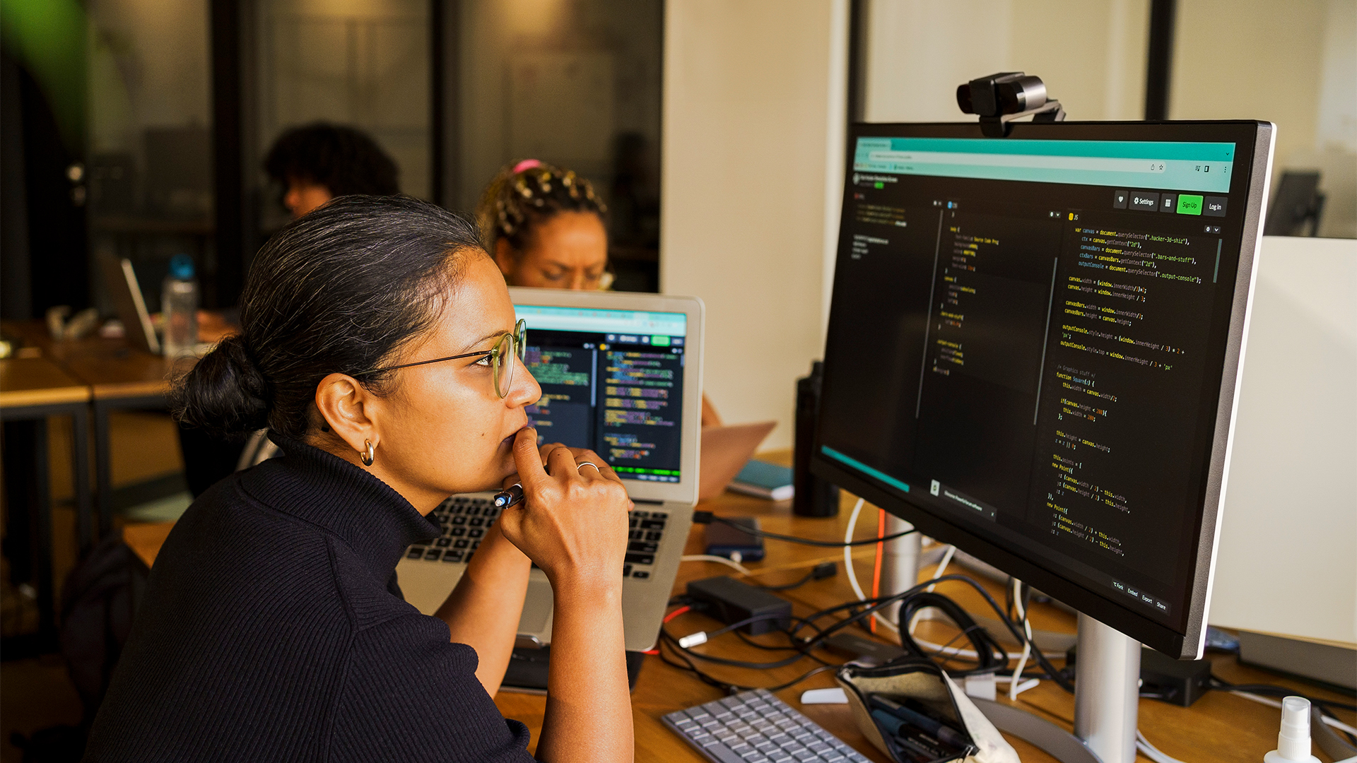 Female software developer looking confused while coding on a desktop computer in an open plan office space, with colleagues in background.