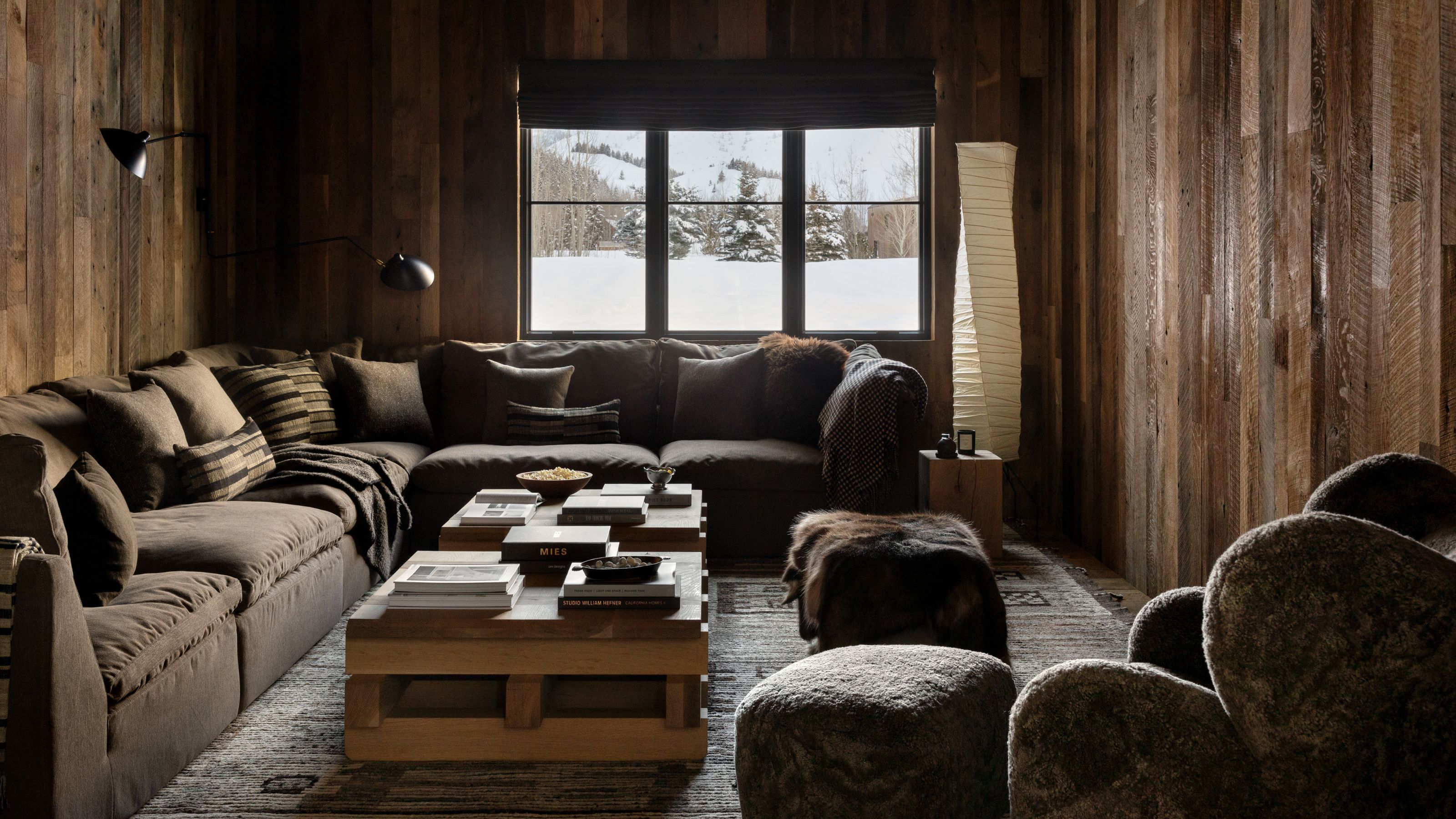 a wood clad media room with a window framing the snowy mountains, a large dark green sectional couch takes up most of the room with a wooden coffee table and shearling arm chair
