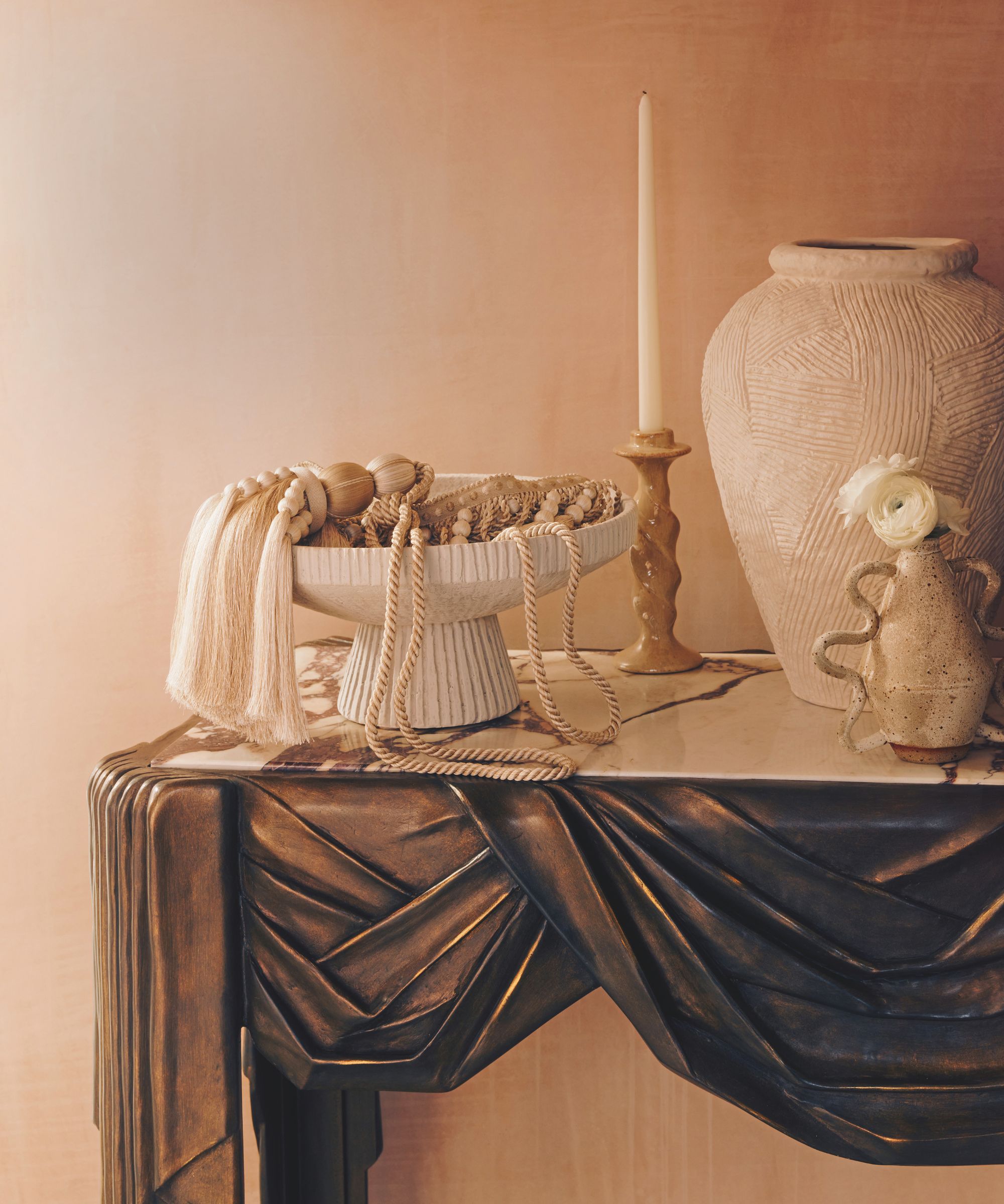 A close-up of a sculpted bronze console table topped with marble. On it sits a pedestal bowl filled with decorative tassels, a tall white textured vase, and a winding yellow taper candle holder.