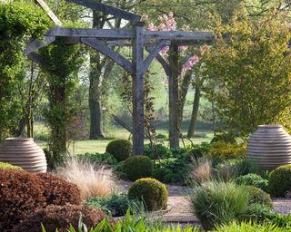 Pergola ideas in a country garden with wood, cedar and timber interspersed with topiary bushes, a selection of trees, long grasses and large stone urns.