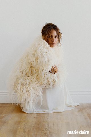 Halle Berry in a white silk dress and feather cape, crouching on a wooden floor against a white wall