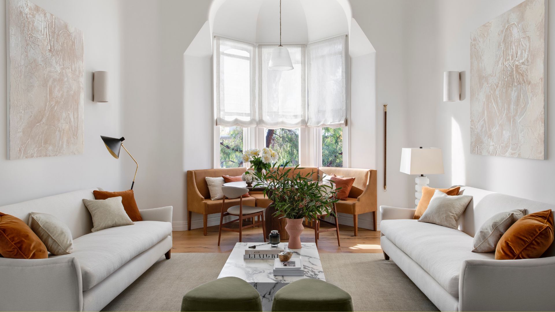 A bright white living room with symmetry in design through white couches, tan and white pillows, hanging wall art, tubular wall sconces, a marble side table with a vase of stems, a stack of books, and a scented candle, beside green stools, and a dining nook by the windows