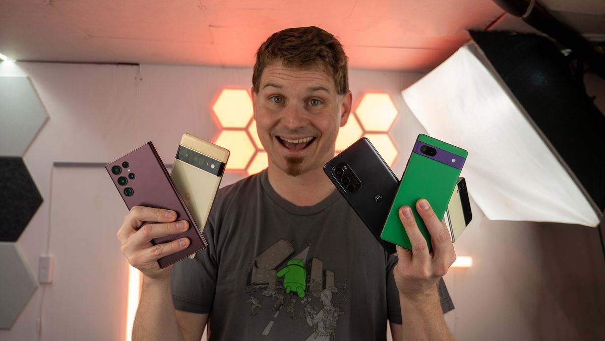 Smiling while holding a Pixel 6a, Pixel 6 Pro, Galaxy S22 Ultra, Moto Edge (2022), and a Galaxy Z Flip 3