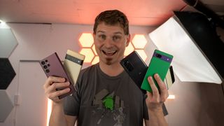 Smiling while holding a Pixel 6a, Pixel 6 Pro, Galaxy S22 Ultra, Moto Edge (2022), and a Galaxy Z Flip 3