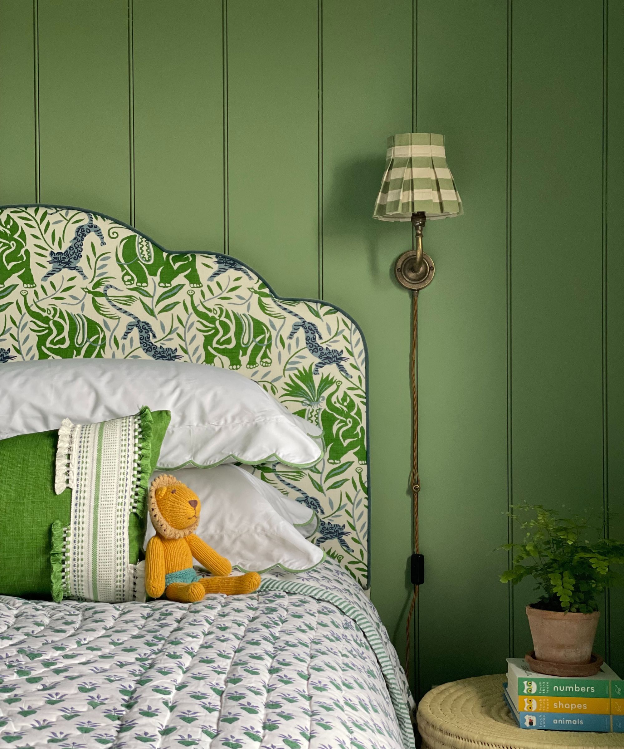 Bedroom with green painted panelled walls, upholstered headboard with floral throw, rattan side table and pleated scocne