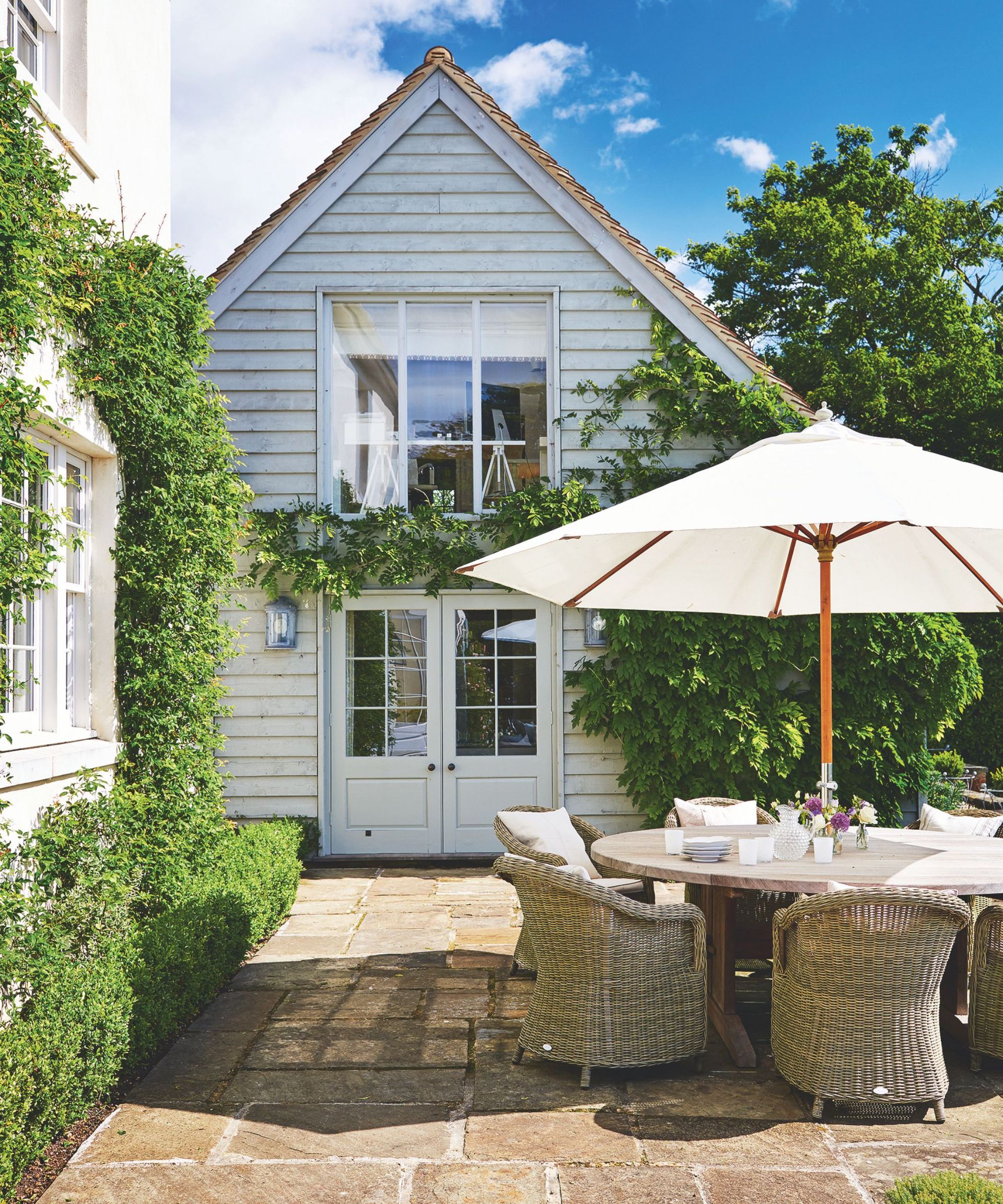 A sunny paved patio with a white panelled house, circular table with wicker chairs and a large white, open umbrella