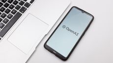 OpenAI logo and symbol pictured on a smartphone screen with smartphone placed on desk next to an Apple MacBook. 