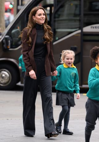 Kate Middleton walking on a sidewalk wearing a brown blazer and gray pants holding hands with a little girl in a teal sweatshirt and gray school uniform skirt.