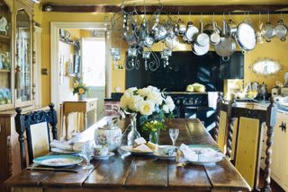 yellow dining room with wooden table and chairs and flowers in centre in kitchen with hanging pans and aga