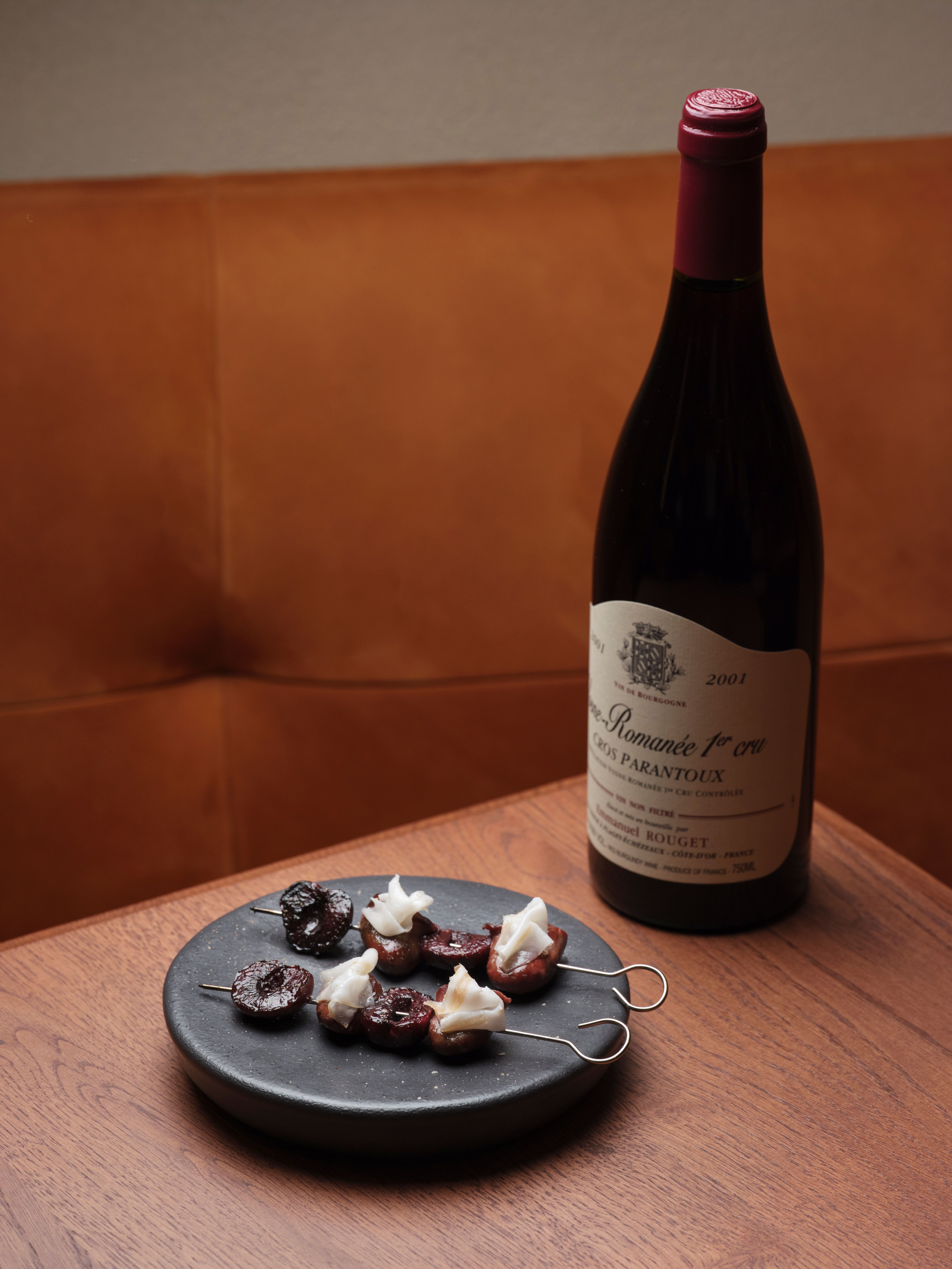 A white ceramic plate with seabass crudo, sprinkled with oil, baslamic vinegar, and spring onions, sits atop a white and black marble top, while in another image, a bottle of red wine sits on a wooden table next a black ceramic snack plate topped with meat, cherry, and cheese skewers.