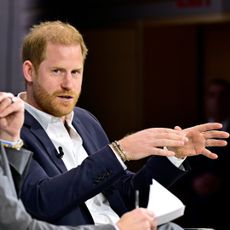 Prince Harry wearing a navy blazer and white shirt speaking to a man in a gray suit whose back is turned to the camera