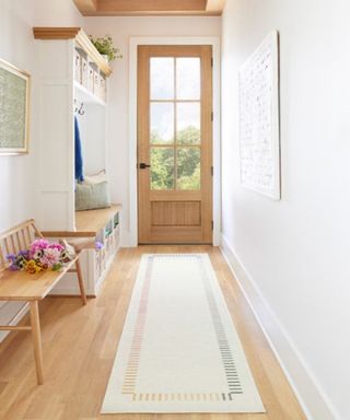 A white entryway with a wooden floor with a white and rainbow rug, a brown door, and a wooden bench