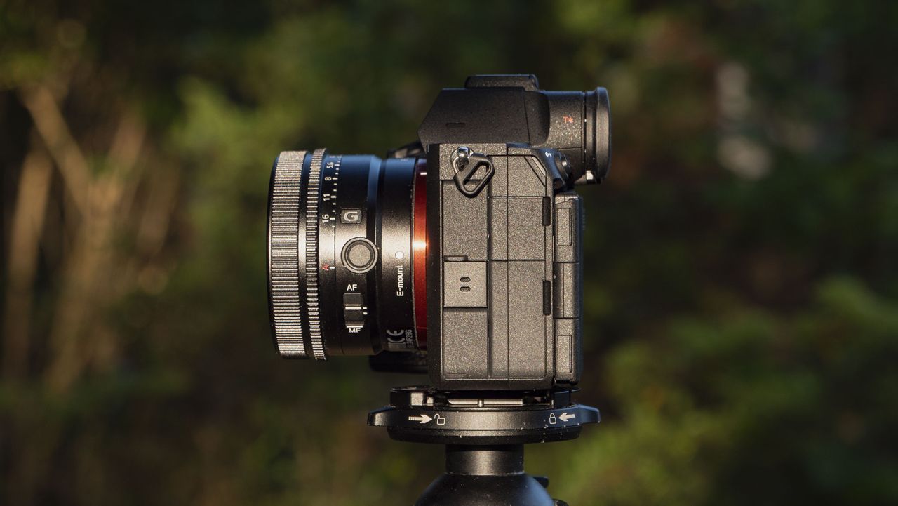 The Sony A7R VI camera with 24mm f/2.8 lens attached, it's mounted to a tripod outdoors and we see the side profile of the connection port doors, there's a natural background