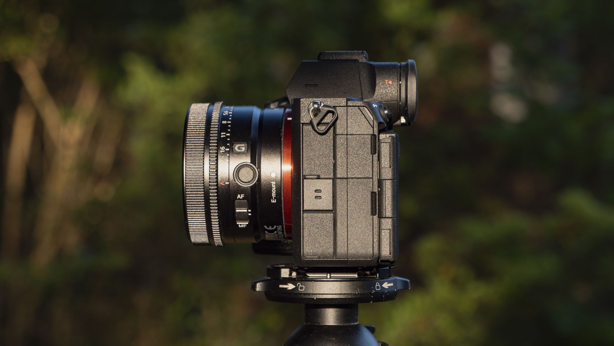 The Sony A7R VI camera with 24mm f/2.8 lens attached, it's mounted to a tripod outdoors and we see the side profile of the connection port doors, there's a natural background